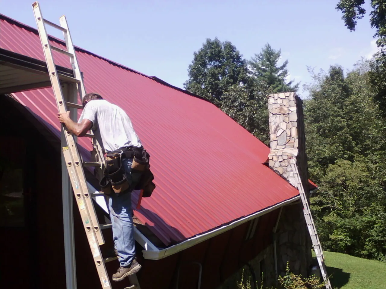 Roofer on a ladder beside a red metal roof and stone chimney — Soffit & Fascia Repair by Roofing Brothers in Red Feather Lakes